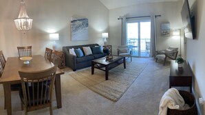 A spacious living room with a dining area, a chandelier, a large window with a view, and a flat-screen TV mounted on the wall. - Inviting and Relaxing Beach and Golf Condo (North Myrtle Beach)