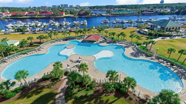 A large, irregularly shaped swimming pool surrounded by palm trees and a marina with boats. - Inviting and Relaxing Beach and Golf Condo (North Myrtle Beach)