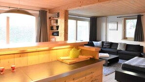 Un salon chaleureux avec un plafond en bois, un canapé, une table basse et un coin cuisine doté d’un plan de travail et d’une bouilloire.