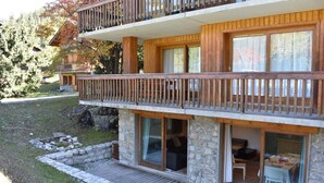 Une maison en bois à deux étages, dotée d’un balcon, de fondations en pierre et d’un petit espace extérieur aménagé pour s’asseoir.