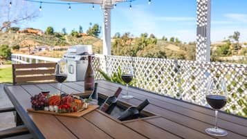 Une table en bois, sur laquelle sont disposés une planche de charcuterie, des bouteilles de vin et des verres, se trouve sur une terrasse extérieure agrémentée d’une clôture en treillis blanc et offrant une vue panoramique.