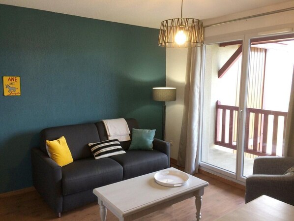 Un salon avec un canapé foncé, une table basse blanche et une lampe. Il y a un panneau accroché au mur et on aperçoit un balcon avec des balustrades en bois.