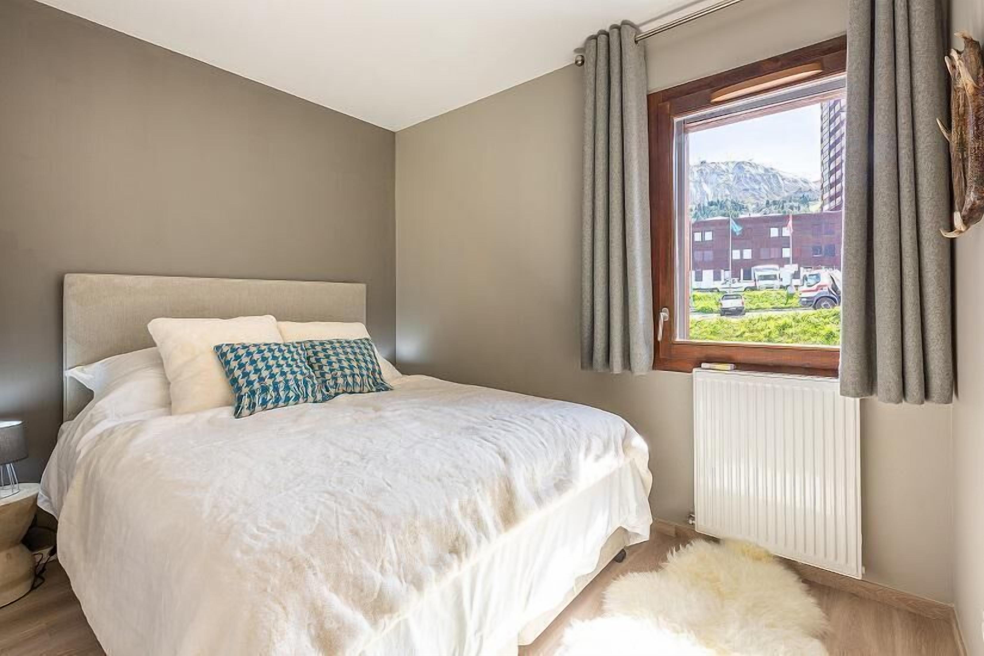 A bedroom with a bed, a window with curtains, a radiator, and a view of buildings and mountains.