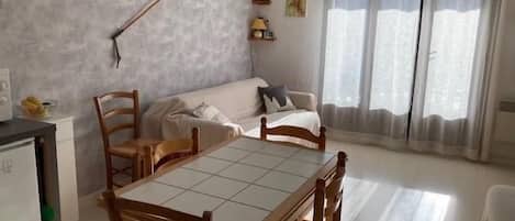 En kompakt opholdsstue med spisebord, stole, sofa, et tekøkken og et vindue med gardiner.