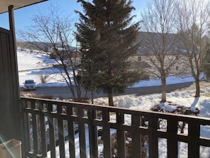 A view from a balcony with a wooden railing, overlooking a snowy landscape with trees and a parked car.