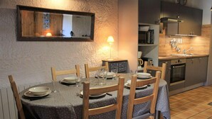 Un coin repas avec une table ronde dressée pour quatre personnes, entourée de chaises en bois. Un miroir est accroché au mur au-dessus de la table, et une lampe se trouve sur la table.