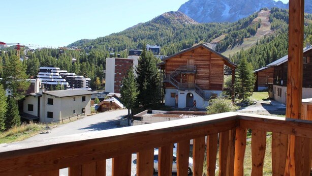 Une cabane en bois dotée d’un balcon, entourée d’un paysage montagneux et de bâtiments modernes.