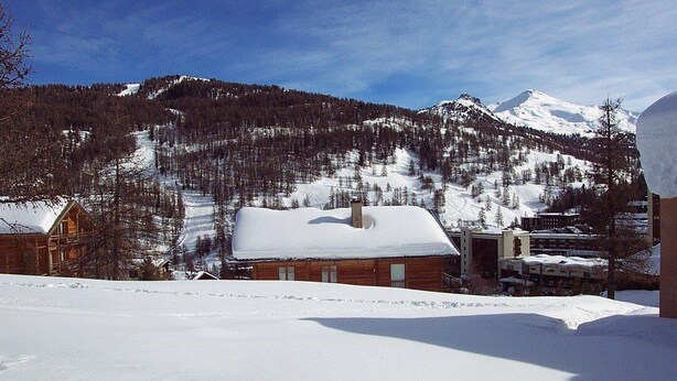 Paisaje montañoso nevado con una cabaña de madera y un edificio con tejado cubierto de nieve.