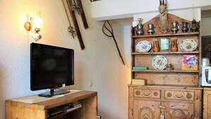 Una habitación con un escritorio de madera, un televisor, una mesa de comedor con sillas y una estantería con objetos decorativos.