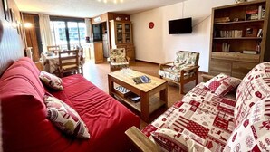 A cozy living room with a red sofa, a wooden coffee table, and a patterned armchair. A television is mounted on the wall, and a bookshelf is visible in the background. - 100m from ski slopes, 2nd floor, balcony, tv, 65m², Vars (Vars)