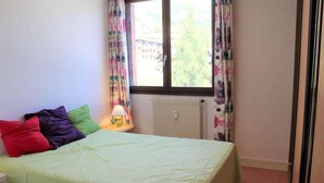 A bedroom with a bed, colorful pillows, a nightstand with a lamp, a window with floral curtains, and a radiator. - 100m from ski slopes, 2nd floor, balcony, tv, 65m², Vars (Vars)
