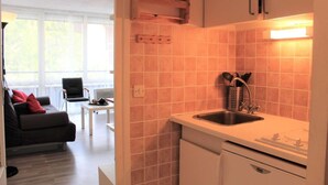 A compact kitchen with a sink, stove, and cabinets. There is a dining area with a chair and table, and a sofa with red cushions.