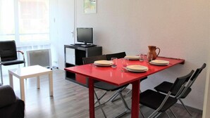 A dining area with a red table set for four, black chairs, a white table, a black chair, a television on a black cabinet, and a painting on the wall.