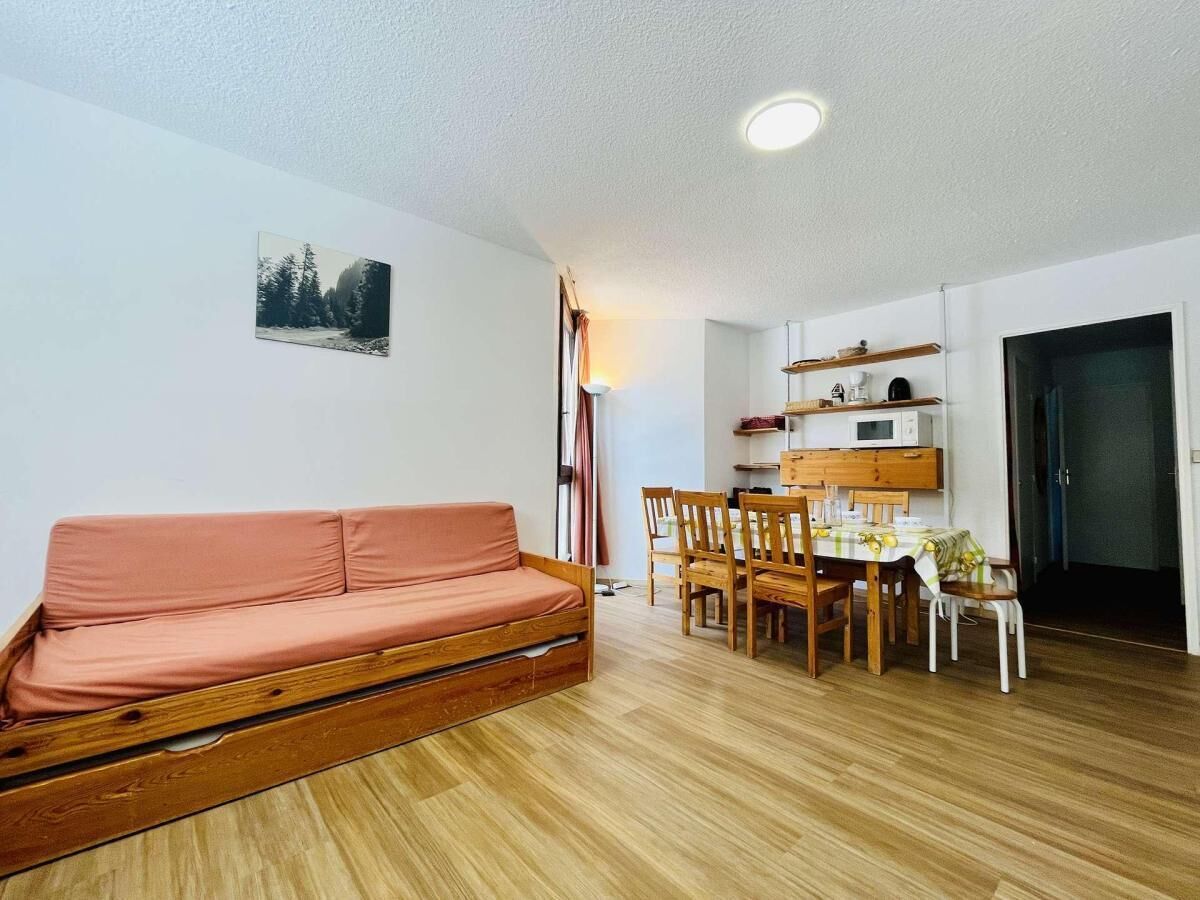 A cozy living room with a wooden sofa, a dining table with chairs, and a kitchen area with shelves and a microwave.
