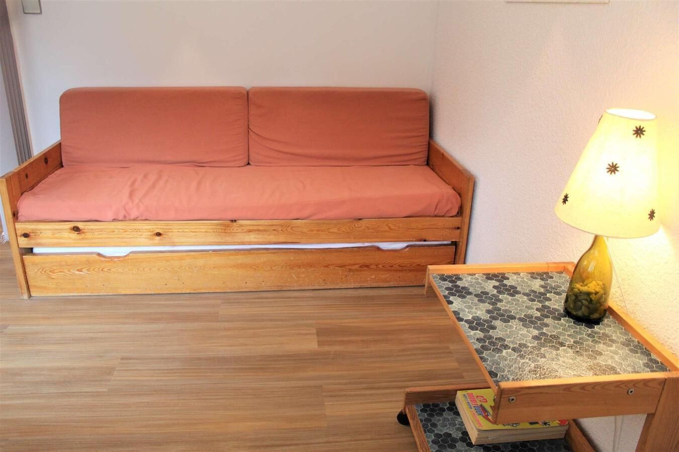 Une petite pièce chaleureuse avec un lit en bois, une petite table sur laquelle se trouve une lampe, et un sol carrelé.
