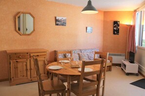 Uma sala de estar aconchegante com uma mesa de jantar de madeira posta para quatro pessoas, um sofá, uma TV em um suporte e uma janela grande com cortinas.