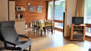 En matplats med träpanel på väggarna, ett matbord med stolar, en tv på ett tv-bord och ett fönster med gardiner.