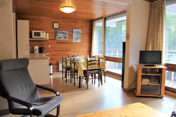 A dining area with wooden walls, a dining table with chairs, a television on a stand, and a window with curtains.