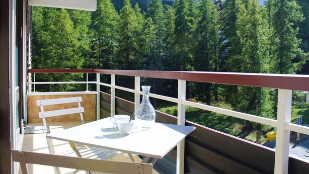 En balkong med ett bord och stolar, en vit vas och utsikt över bergen.