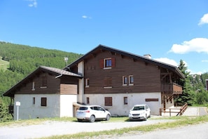 Un edificio in legno a due piani con un balcone, auto parcheggiate di fronte e montagne sullo sfondo.