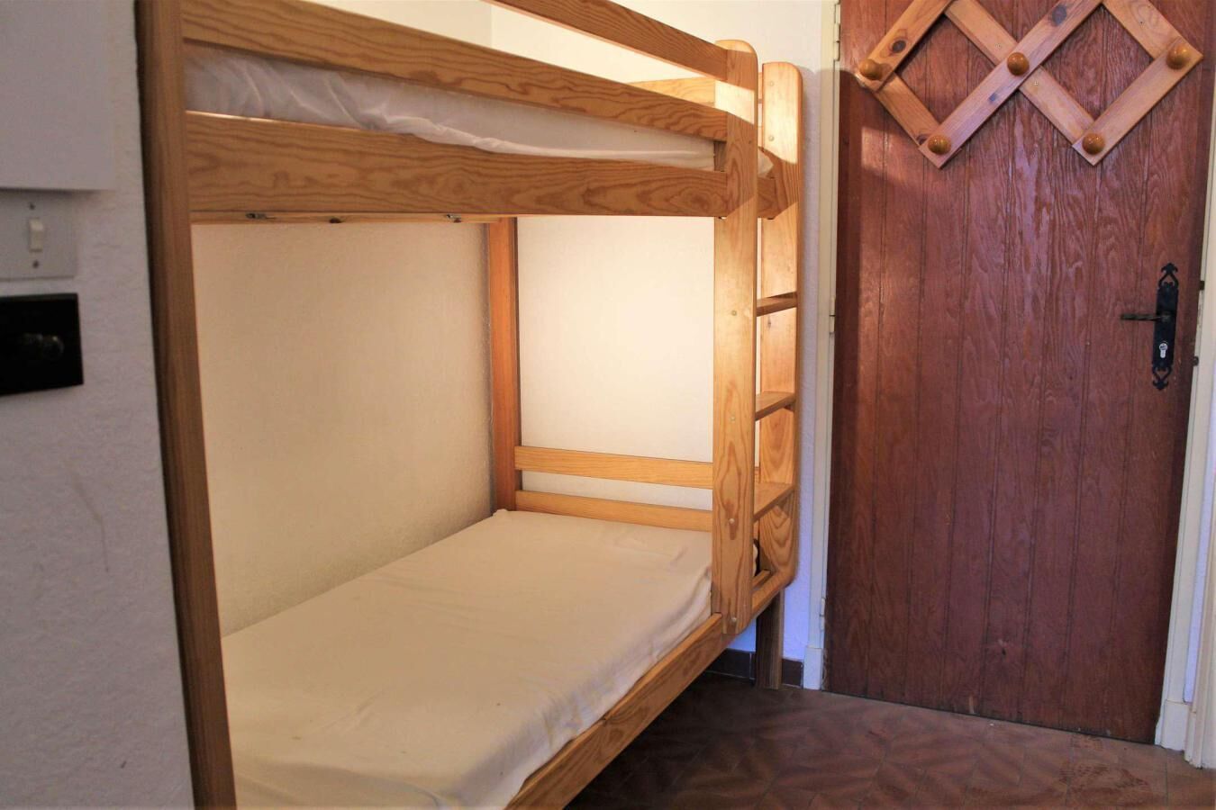 Una cama litera con estructura de madera y colchón, una puerta de madera con diseño de rombos y un enchufe de pared.