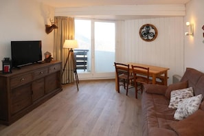 A cozy living room with a wooden TV stand, a table with chairs, a sofa, and a large window with curtains.