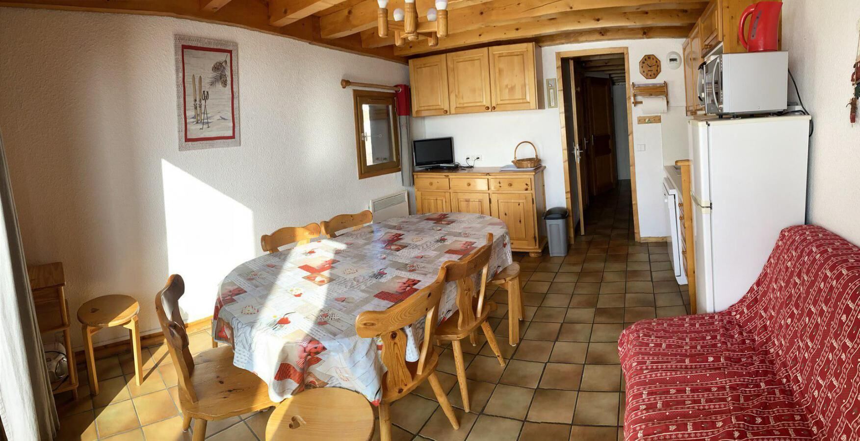 A cozy kitchen with wooden cabinets, a dining table with chairs, a red patterned sofa, and a tiled floor.