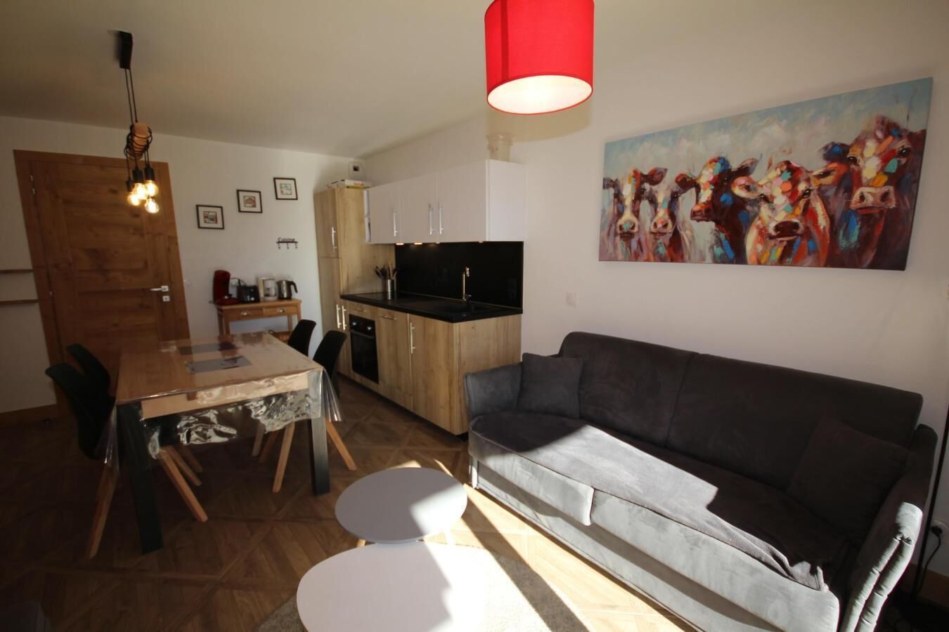 Un salon moderne avec un canapé gris, une table à manger avec des chaises, un coin cuisine avec des placards et un téléviseur, ainsi qu’un luminaire suspendu rouge.
