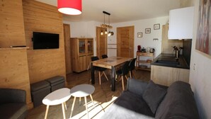 A compact living space with a wooden interior, a dining area with a table and chairs, a sofa, a kitchen with a sink and countertop, and a TV mounted on the wall. - 100m from ski slopes, Ground floor, terrace, tv, ski locker, 47m², Les Saisies (Hauteluce)