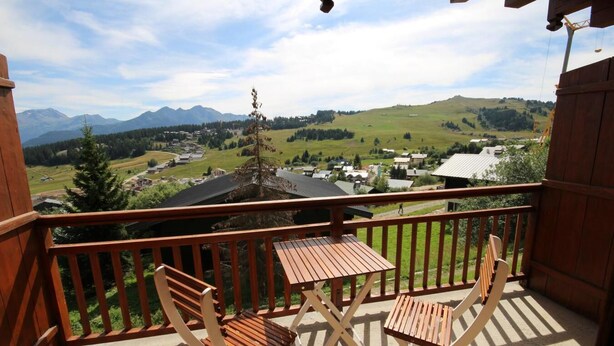 Un balcon en bois, agrémenté d’une table et de chaises, d’où l’on peut admirer un village et des montagnes.