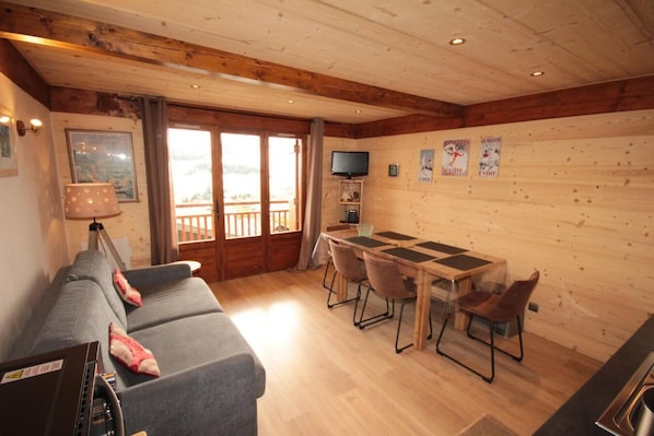A cozy room with a sofa, a dining table with chairs, a TV, and a balcony with a railing.