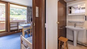 A room with a wooden door, a window with a view of greenery, a small table with a chair, and a bathroom with a sink and mirror. - 5m from ski slopes, 1st floor, view ski slopes, balcony, tv, 21m², La Plagne (La Plagne)