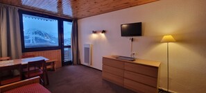 Ein Zimmer mit Holzboden, einem an der Wand befestigten Fernseher, einem Schreibtisch mit Stuhl, einer Lampe und Blick auf verschneite Berge durch ein Fenster.