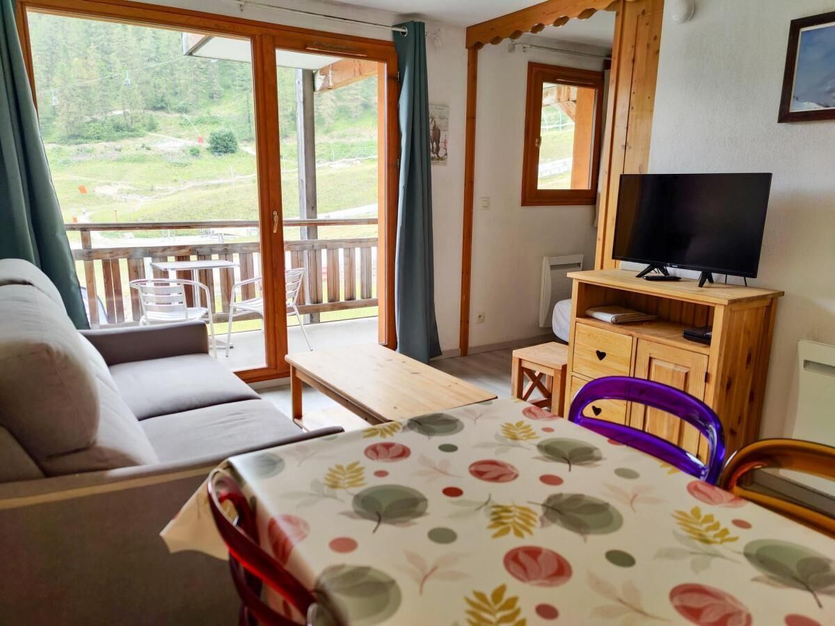 A room with a sofa, a dining table with a floral tablecloth, a television on a wooden stand, and a view of a balcony with outdoor furniture.