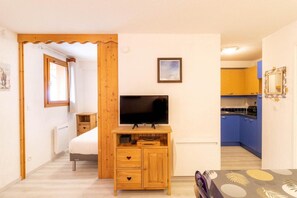 A compact room with a wooden TV stand, a bed, a small table, and a kitchenette with blue cabinets. - 100m from resort center, Ground floor, swimming pool, sauna, balcony, tv, ski locker, 33m² (Les Orres)