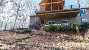 A two-story house with a wooden balcony, a staircase leading to the entrance, and a view of leaf-covered ground. - The View on Lake Lanier lake view & dock. (Gainesville)