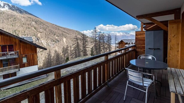 A wooden balcony with a table and chairs, overlooking a mountainous landscape.