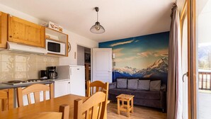 A cozy kitchen with wooden cabinets, a dining table, and chairs. There's a microwave, stove, and refrigerator. A mural of mountains is on the wall, and a sliding glass door leads to an outdoor area.