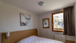 A bedroom with a wooden headboard, a bed with a white quilt, a wall-mounted lamp, two framed pictures, a window with curtains, and a radiator.