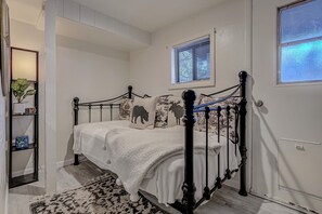 A bedroom with a bed, a window with blinds, a shelf with plants, and a door.
