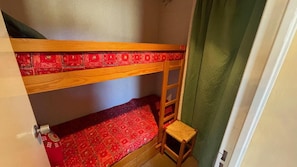 Uma cama beliche com colcha estampada vermelha, uma escada de madeira e um banquinho de madeira.