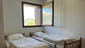 Quarto com duas camas de solteiro, janela com vista para montanhas e cabeceira de madeira.