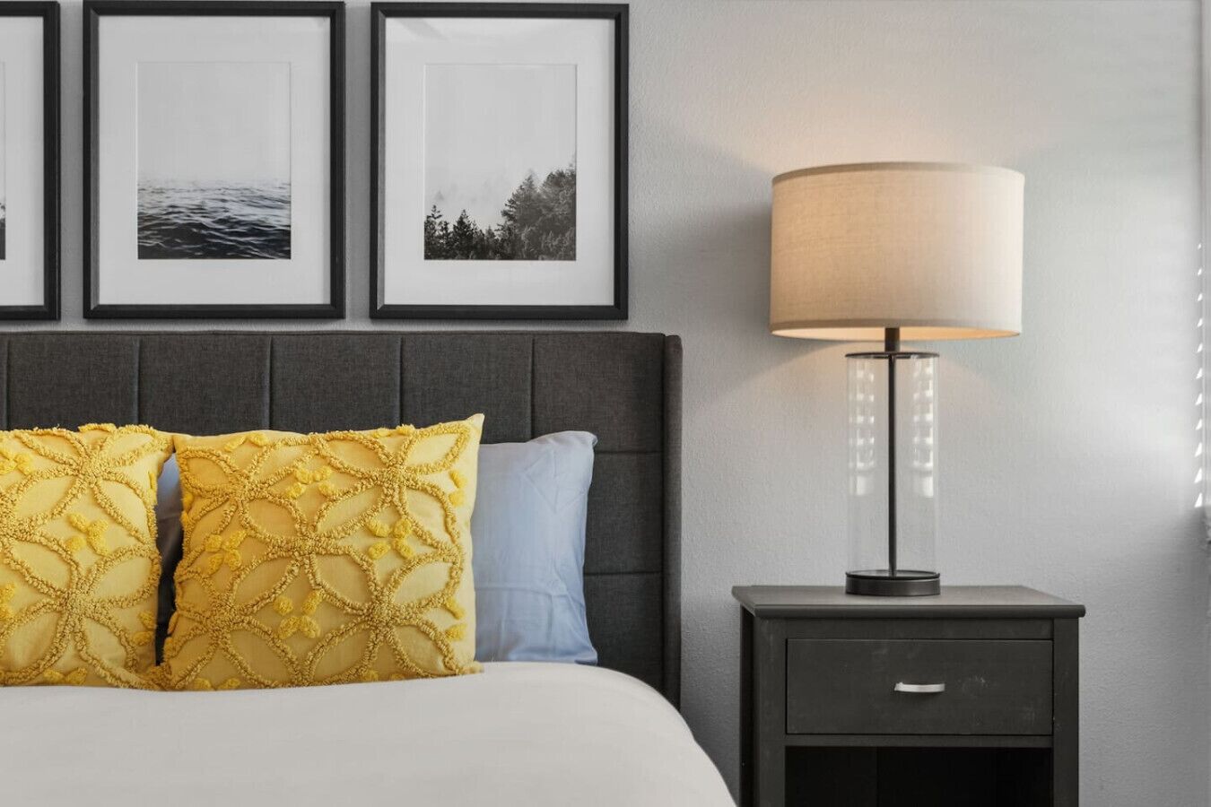 Habitación de hotel ordenada con cama, dos almohadas, mesita de noche con lámpara y cuadros en la pared.