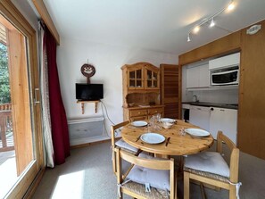 A dining area with a round wooden table, cushioned chairs, a television, and a kitchen with a microwave.