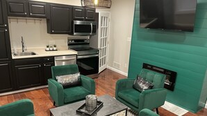 Une cuisine moderne avec des meubles de couleur sombre, un four à micro-ondes, un four et un lave-vaisselle. On y trouve deux fauteuils verts ornés de coussins décoratifs et une table basse noire. Un téléviseur à écran plat est fixé sur un mur d’accent de couleur turquoise.
