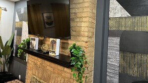 Un salon moderne avec un téléviseur à écran plat fixé à un mur en briques, une étagère en bois foncé ornée d’objets décoratifs et des plantes vertes.