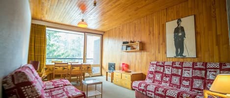 Um quarto acolhedor com teto de madeira, um sofá com manta estampada, uma mesa com cadeiras e uma grande janela com cortinas.