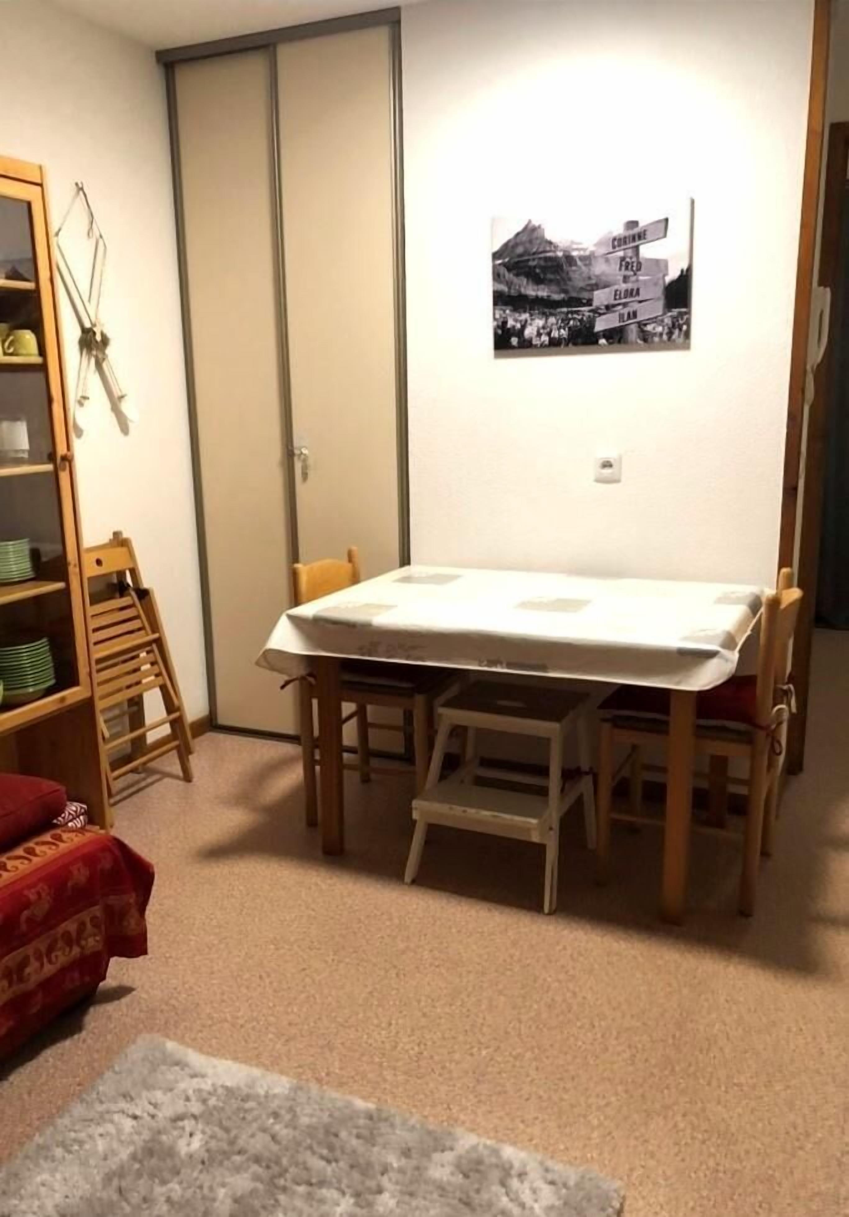 A small dining area with a table and chairs, a wooden chair with a red cushion, a shelf with dishes, and a framed picture on the wall.