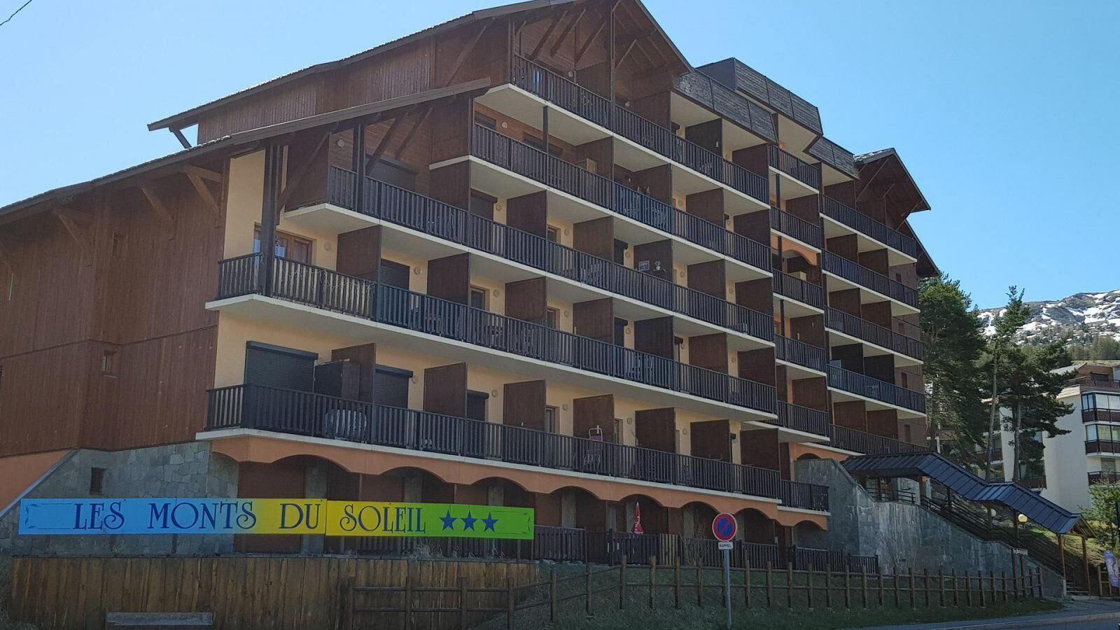 Um hotel com vários andares, varandas, uma placa com a inscrição "LES MONTS DU SOLEIL" e uma pista de esqui ao fundo.