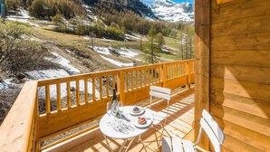 A wooden deck with a table and chairs, overlooking a mountainous landscape with snow patches.